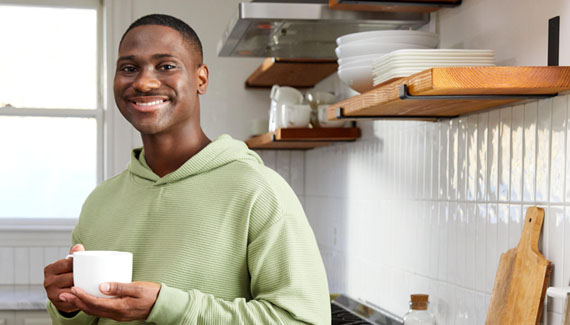 Man standing in his kitchen holding a cup of coffee smiling