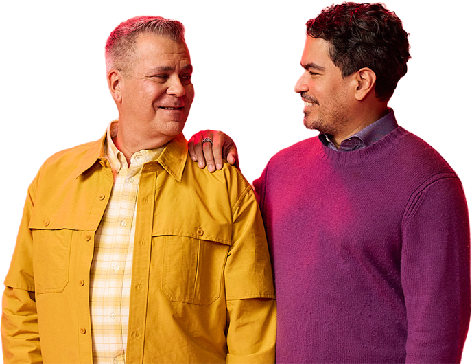 A white male in his fifties and latino man in his thirties smile at each other, his hand resting on the shoulder of the other. The older wearing a yellow jacket the other a purple sweater
