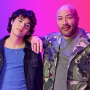 Two individuals pose against a pink background. One leans casually, wearing a blue shirt and dark jacket, and the other smiles in a camo jacket and baseball cap, giving a relaxed vibe.
