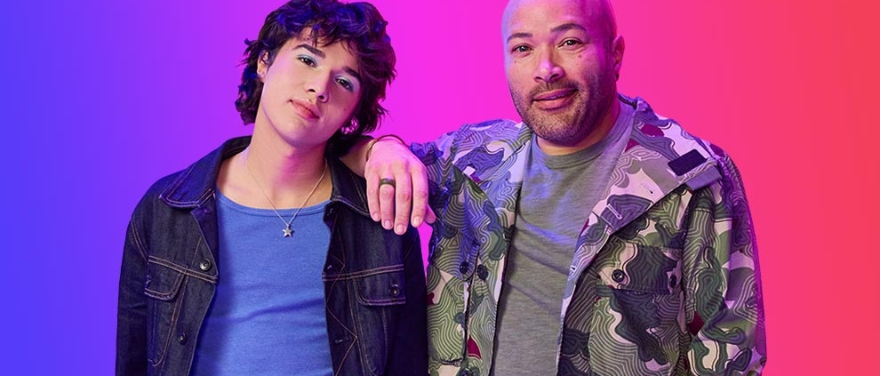 Two individuals pose against a pink background. One leans casually, wearing a blue shirt and dark jacket, and the other smiles in a camo jacket and baseball cap, giving a relaxed vibe.