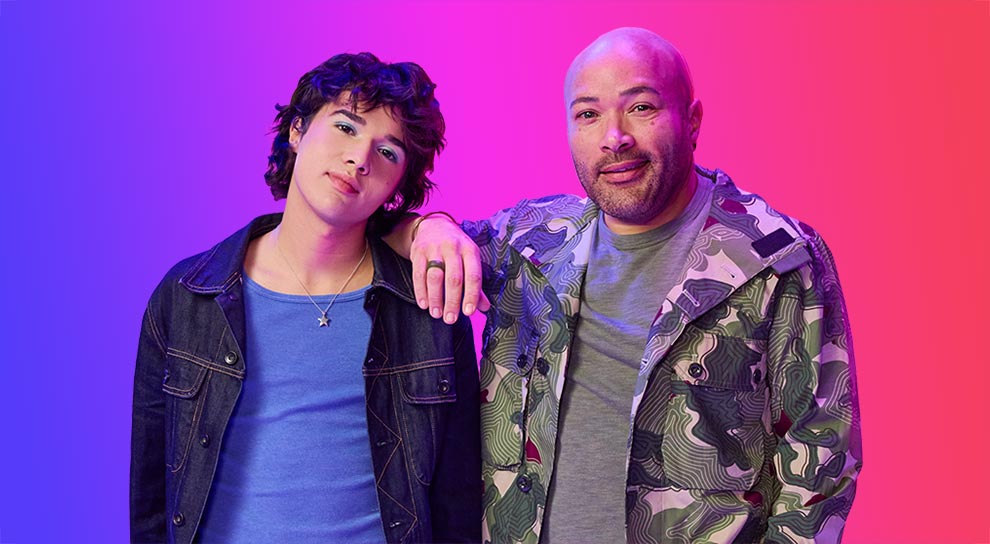 Two individuals pose against a pink background. One leans casually, wearing a blue shirt and dark jacket, and the other smiles in a camo jacket and baseball cap, giving a relaxed vibe.