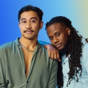 An Asian male and black male against a vibrant blue and yellow gradient backdrop. One leans on the other's shoulder, both projecting a calm, relaxed vibe.