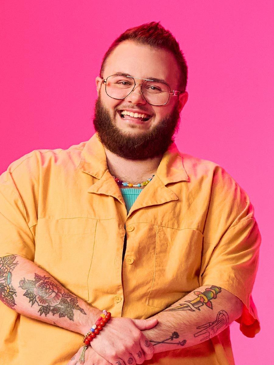 A heavy-set man with tattoos, glasses, and a beard smiles while wearing an yellow shirt and pants against a vibrant pink background, creating a cheerful mood.