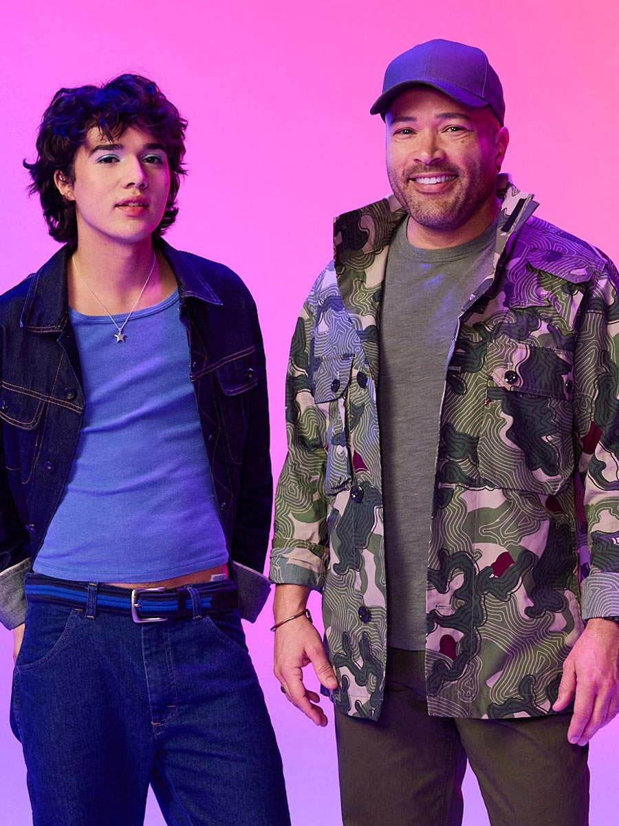 Two individuals pose against a pink background. One leans casually, wearing a blue shirt and dark jacket, and the other smiles in a camo jacket and baseball cap, giving a relaxed vibe.