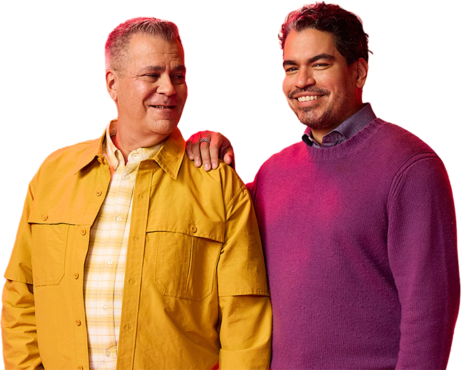 A couple smiling at each other, standing against a colorful background. One wears a yellow jacket; the other dons a purple sweater. Warm, friendly atmosphere.