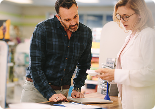 A pharmacist compares medication with a man in a plaid shirt at a pharmacy counter