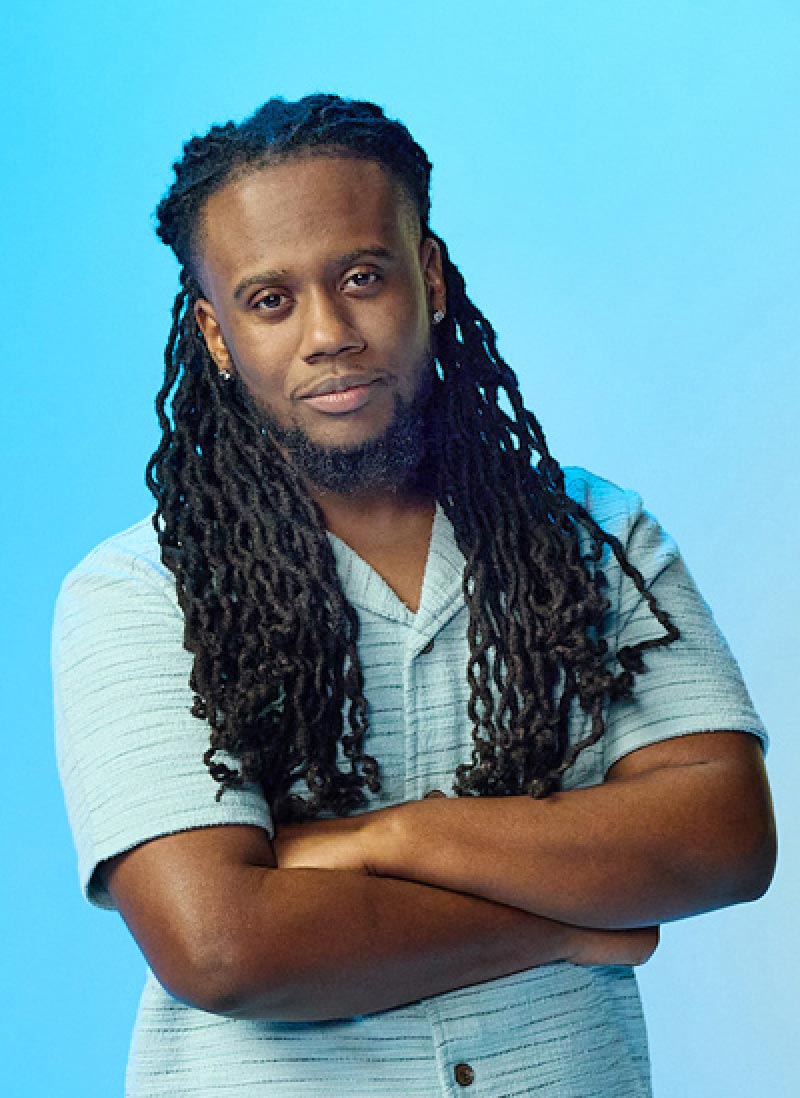 A man with long locks and a beard smiles confidently with crossed arms. They wear a light blue short-sleeve shirt against a bright blue background.