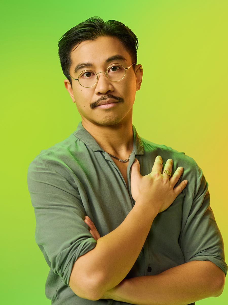 An Asian male in his early thirties wearing glasses and a gray shirt poses against a vibrant yellow-green background, hand on chest. The mood is confident and thoughtful.
