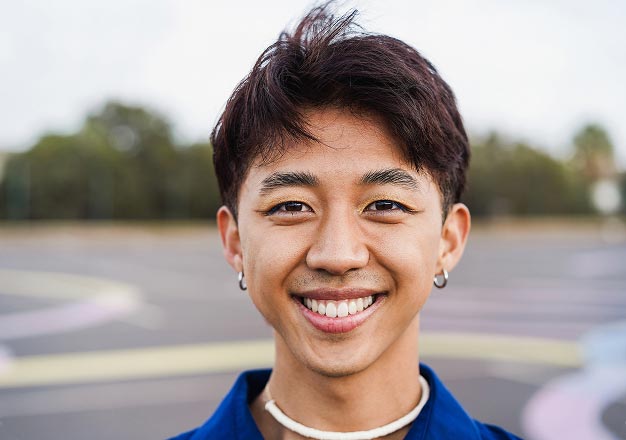 Smiling young Asian male with short dark hair, wearing hoop earrings and a necklace, outdoors. Blurred background of trees and pavement.