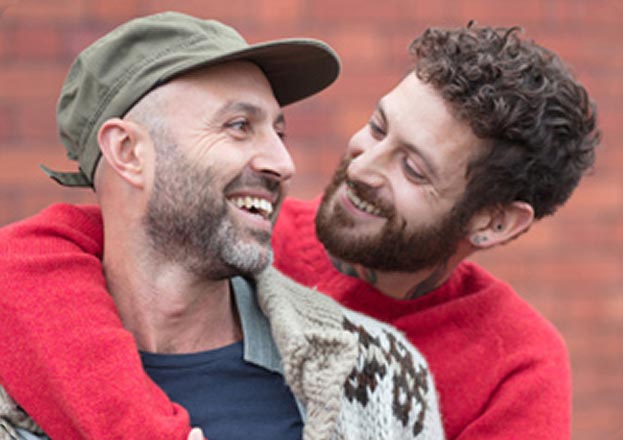 Two smiling people share a warm embrace against a brick wall. One wears a red sweater, the other sports a cap and patterned sweater, exuding joy and affection.