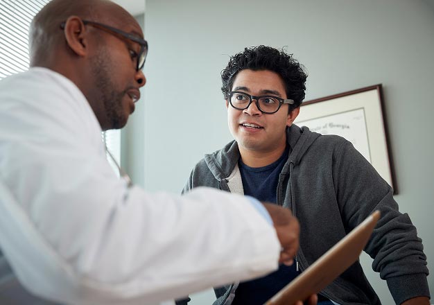 A black male doctor in a white coat speaks with a bi-racial patient wearing glasses and a hoodie while looking at a tablet. The setting is professional yet relaxed, suggesting attentive communication.