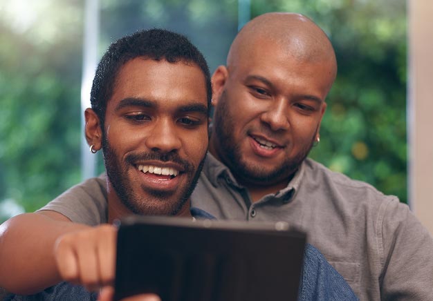 A latino man and black man are sitting close together, smiling while looking at a tablet. The background features greenery, suggesting an indoor setting with natural light. One man has short hair and a beard, while the other has curly hair and wears earrings.