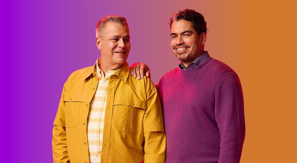A couple smiling at each other, standing against a colorful background. One wears a yellow jacket; the other dons a purple sweater. Warm, friendly atmosphere.